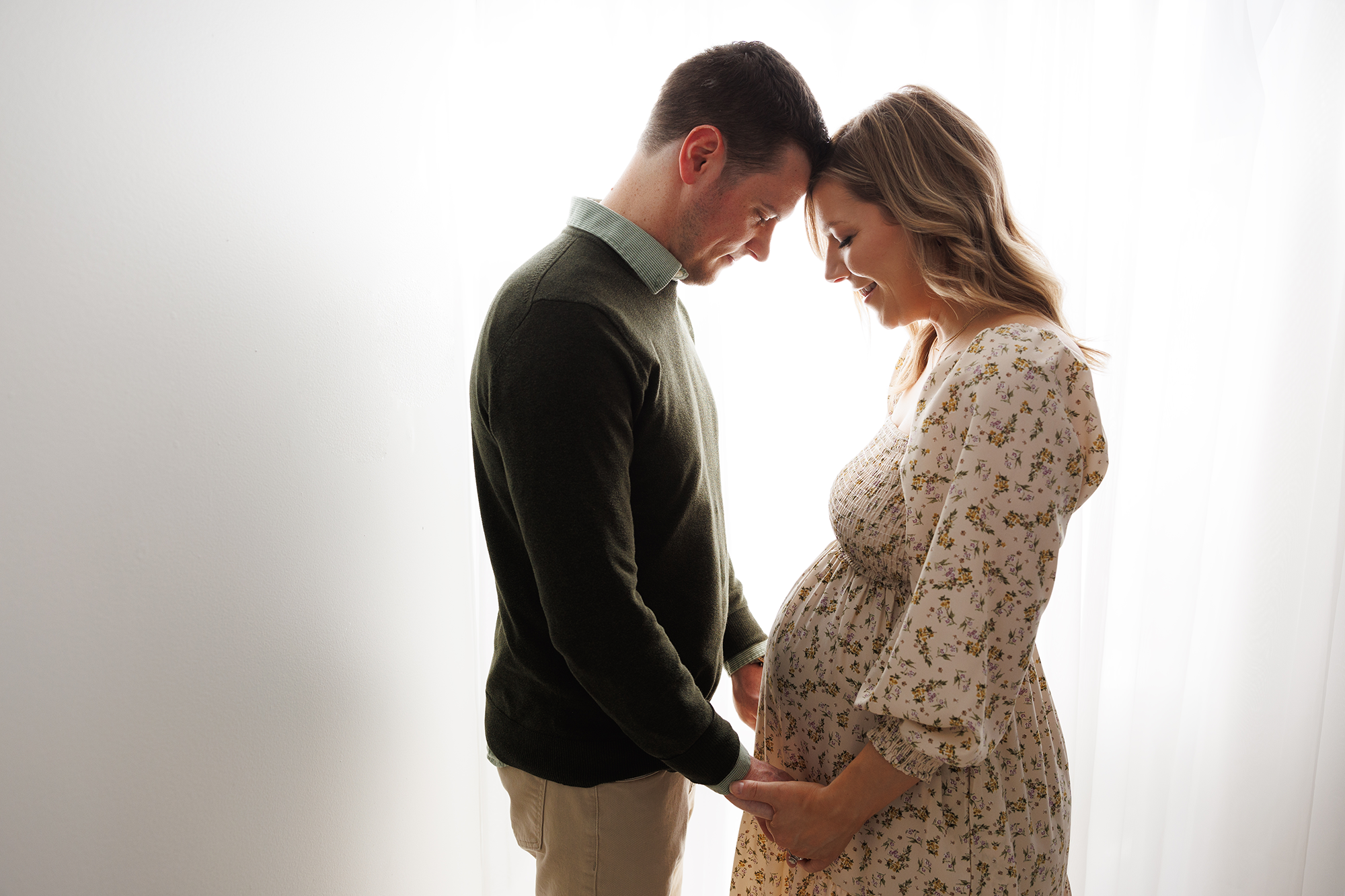 Christine Retzer, St. Charles, IL Newborn Photographer, photo of a couple in front of window holding hands looking at Mom's pregnant belly.